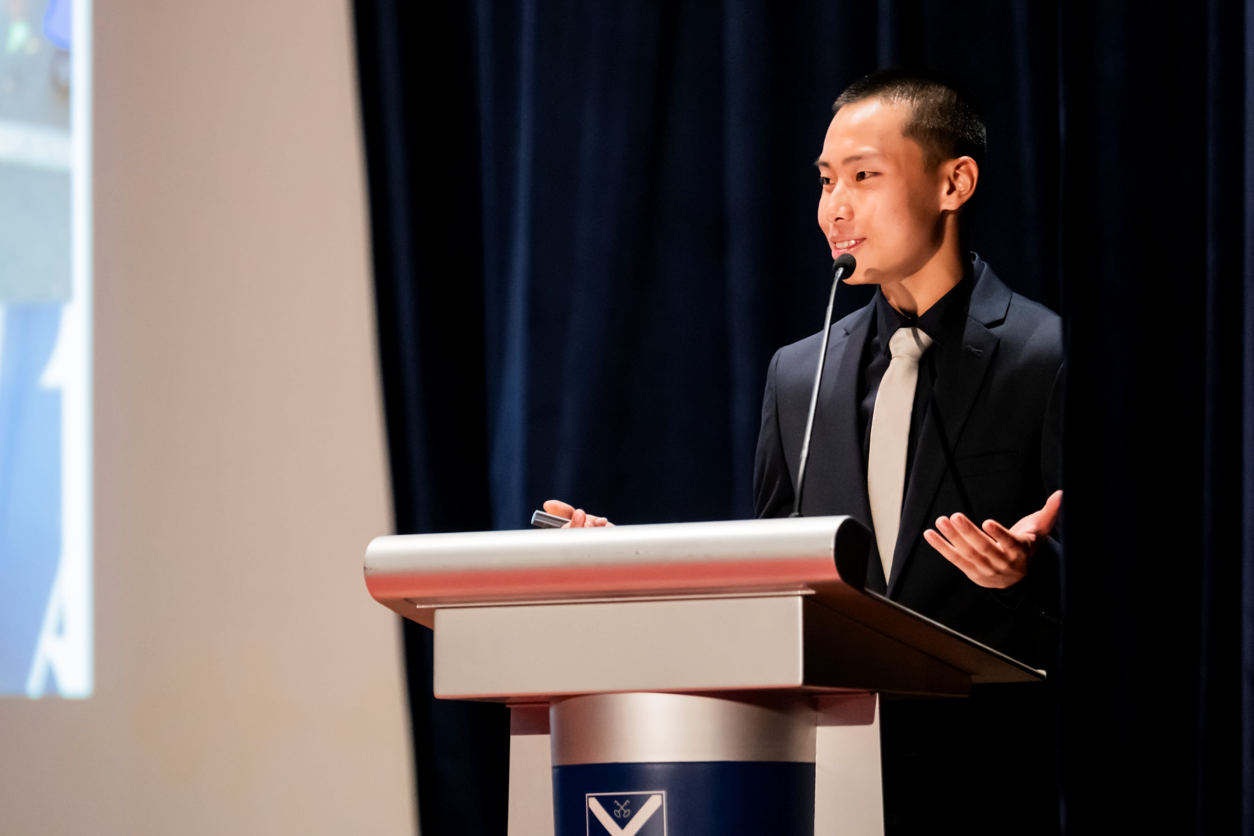 Valedictorian of the Class of 2025, Teo Jin Rui, in a dark suit and light tie speaking at a podium on stage with open hand gestures.